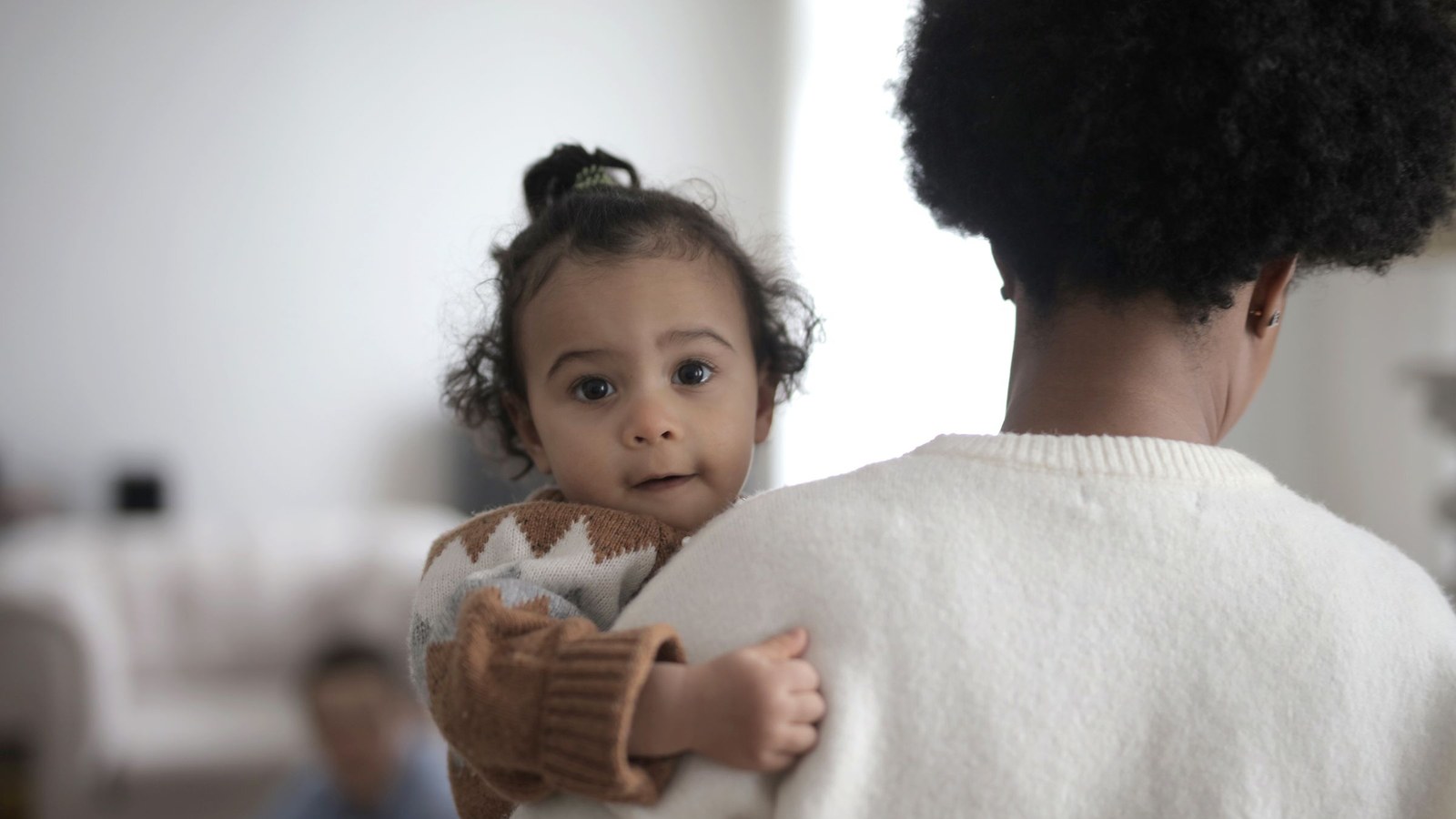 A toddler in a knit sweater held in one parent's arms at home, leaning into their shoulder and holding on